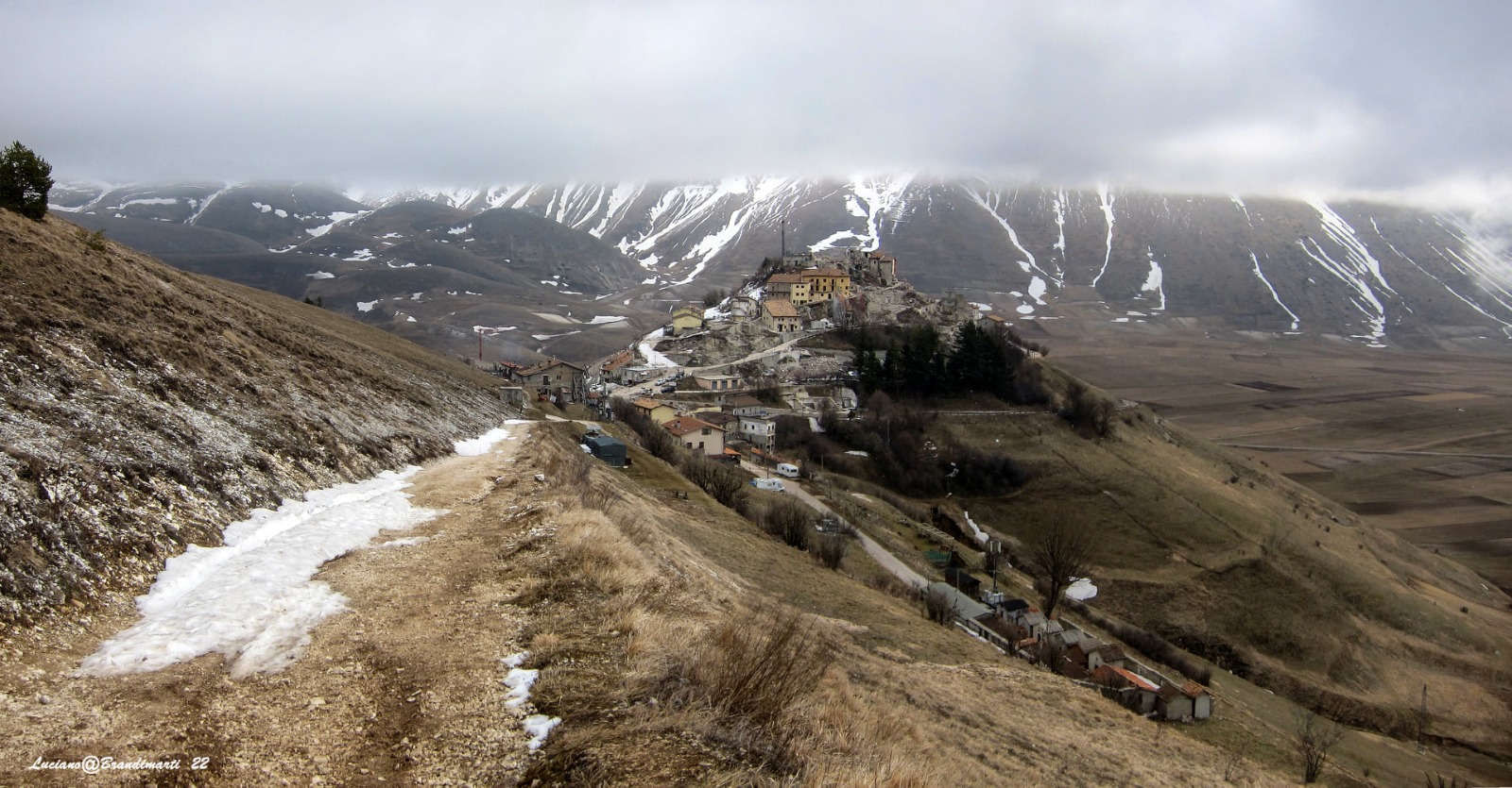 Front view of a partially snow-covered path leading to Castelluccio di Norcia, in the centre of the photo. To the right of the hill where the village stands, part of the Pian Grande can be glimpsed. In the background, the Sibillini Mountains covered partially in show, are covered by a layer of mist.