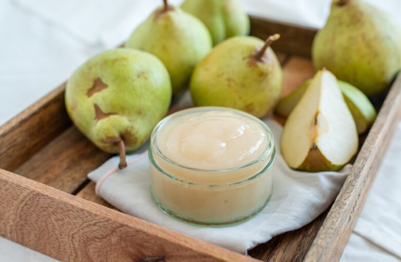 in the foreground of the picture, there is a small glass jar containing Monteleone di Orvieto pear jam. In the blurred background, there are pears, either whole or cut in half. The whole is resting on a wooden tray with raised edges.