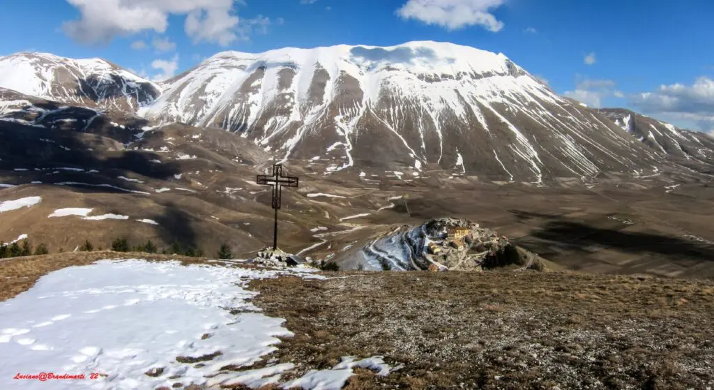immagine panoramica dall’alto di castelluccio di norcia su una collinetta che si staglia in mezzo alla piana di castelluccio di norcia. Le punte dei rilievi dei Monti Sibillini intorno sono innevate.