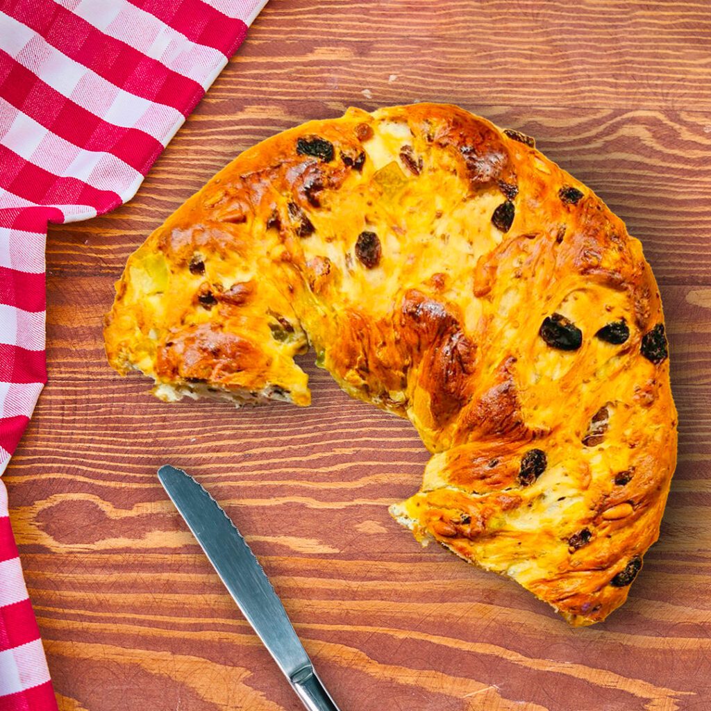 Photo taken from above of a section of torcolo of Saint Constantius. Torcolo is a typical Umbrian cake, specifically from the area of Perugia, shaped like a doughnut with a central hole. In the photo, from above it is possible to see half of this cake with raisins and candied fruit on top. The torcolo is placed on a wooden table, a knife is placed next to the torcolo and on the left side of the picture there is a checked tablecloth.