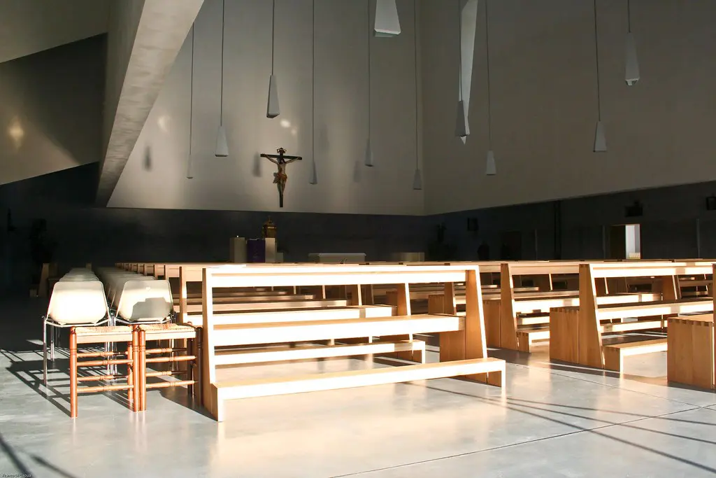 foto della vista interna della chiesa di San Paolo a Foligno. Sulla parte bassa della foto, ci sono due file che si sviluppano in verticale di panche di legno utilizzate per sedersi e pregare all’interno della Chiesa. Accando alla fila di sinistra, due file di sedie affiancano la fila di panche di legno. Le panche sono sovrastate dalla struttura a parallelepipedo interna alla chiesa da cui pendono tanti lampadari tutti uguali, con paralumi dalla forma geometrica irregolare. Sulla parete in fondo è appeso un grande crocifisso in legno.