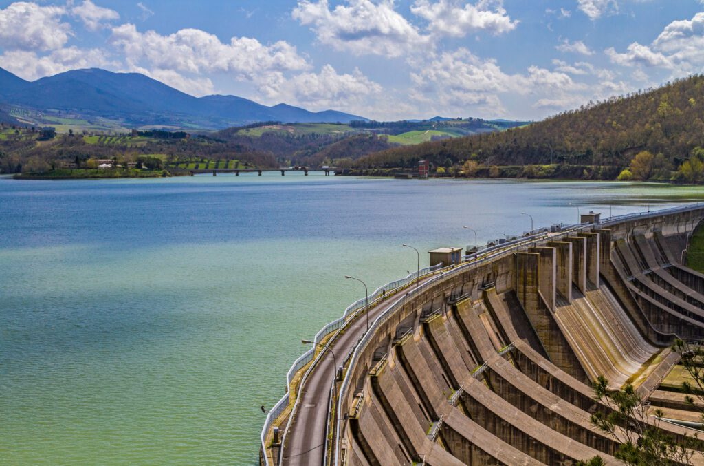 foto ravvicinata del muro di contenimento della diga da cui ha origine il lago di Corbata. In basso a destra, dall’alto è raffigurato l’interno del muro di contenimento, con piloni a intervalli regolari che percorrono il muro e poi discendono verso il basso. Sopra il muro, una strada asfaltata lo percorre per tutta la lunghezza e a un lato, a intervalli regolai, ci sono dei lampioni. Sul lato sinistro in basso dell’immagine, dall’alto si vede l’acqua del lago, mentre sullo sfondo ci sono le montagne che circondano il lago. Il cielo è sereno e con delle nubi.