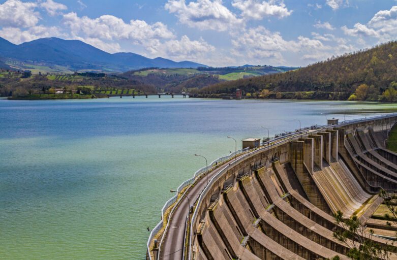 foto ravvicinata del muro di contenimento della diga da cui ha origine il lago di Corbata. In basso a destra, dall’alto è raffigurato l’interno del muro di contenimento, con piloni a intervalli regolari che percorrono il muro e poi discendono verso il basso. Sopra il muro, una strada asfaltata lo percorre per tutta la lunghezza e a un lato, a intervalli regolai, ci sono dei lampioni. Sul lato sinistro in basso dell’immagine, dall’alto si vede l’acqua del lago, mentre sullo sfondo ci sono le montagne che circondano il lago. Il cielo è sereno e con delle nubi.