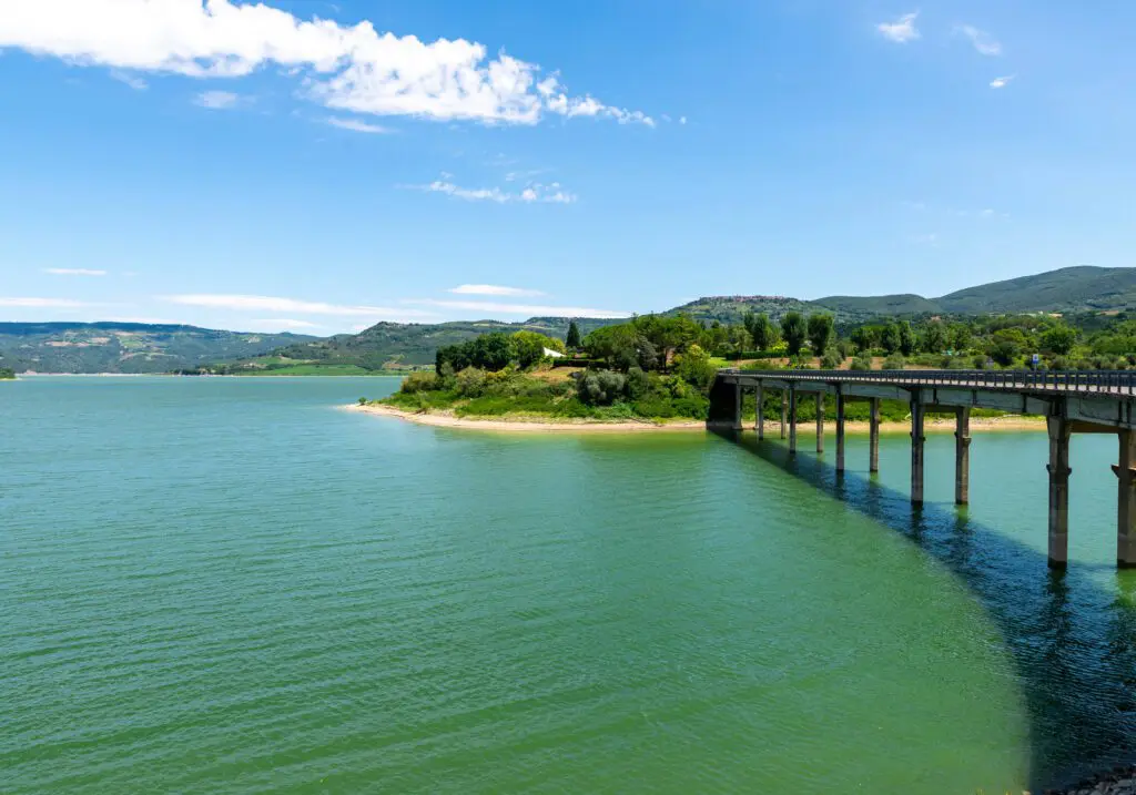 immagine che raffigura da vicino una vista sul Lago di Corbara. Sul lato sinistro dell’immagine, c’è un’inquadratura ravvicinata dell’acqua del lago. Sul lato destro dell’immagine, un ponte attraversa una sezione del lago per raggiungere una riva ricca di vegetazione. Sullo sfondo, le montagne fanno da contorno al lago sotto un cielo sereno con nubi sparse.