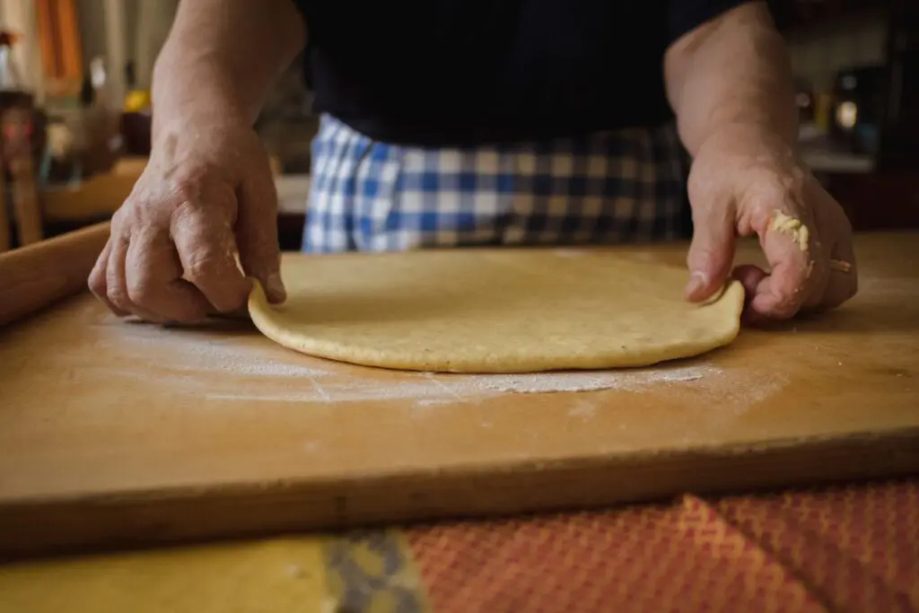 inquadratura ravvicinata frontale delle mani di una signora che tiene l’impasto steso della torta al testo sui bordi laterali. L’impasto è appoggiato su un tagliere di legno e ha la forma tipica della torta al testo rotonda e schiacciata.