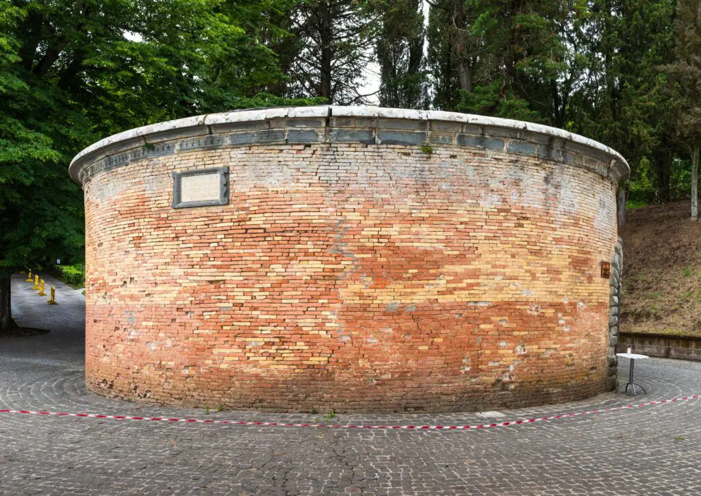 vista esterna ravvicinata della parte esterna del Pozzo di San Patrizio, una struttura circolare composta da mattoncini di tufo con varie sfumature di colori. Piante di varie tipologie fanno da sfondo alla foto e circondano l’area in cui si trova il pozzo.