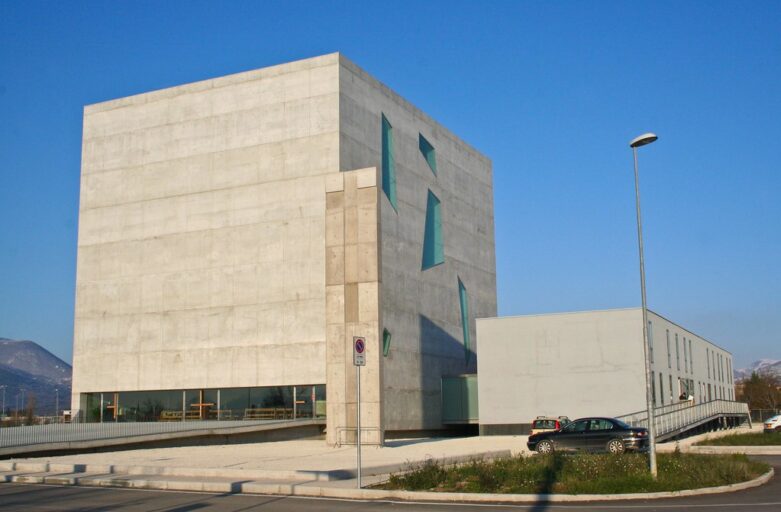 Outsie view of the church of St Paul in Foligno and the surrounding area, designed by architects Massimiliano and Doriana Fuksas. On the left side of the photo, there is the larger building where the church is located inside. On one side, the main facade of the church is made of reinforced concrete, with no openings; a slightly sloping walkway leads to the base of the building, where a long stained-glass window forms the entrance to the church. On the side facade, on the other hand, there are windows of different sizes and shapes. Opposite to the church, to the right of the picture, there is another smaller concrete building, with windows arranged in two rows along the long side, facing outwards. In a small square next to the entrance walkway, in the centre of the image, is a crucifix carved from a 13-metre high block of concrete and white Carrara marble by the artist Enzo Cucchi. In front of the church, there is a car park where two cars are parked.
