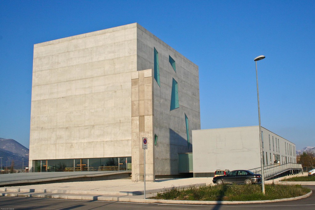 Outsie view of the church of St Paul in Foligno and the surrounding area, designed by architects Massimiliano and Doriana Fuksas. On the left side of the photo, there is the larger building where the church is located inside. On one side, the main facade of the church is made of reinforced concrete, with no openings; a slightly sloping walkway leads to the base of the building, where a long stained-glass window forms the entrance to the church. On the side facade, on the other hand, there are windows of different sizes and shapes. Opposite to the church, to the right of the picture, there is another smaller concrete building, with windows arranged in two rows along the long side, facing outwards. In a small square next to the entrance walkway, in the centre of the image, is a crucifix carved from a 13-metre high block of concrete and white Carrara marble by the artist Enzo Cucchi. In front of the church, there is a car park where two cars are parked.
