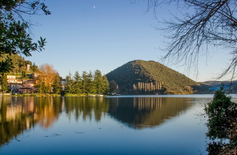 View of Lake Piediluco and the mountains surrounding it. On the left bank of the picture, a group of houses is illuminated by the sun rays and, next to it, there is a small group of pine trees.