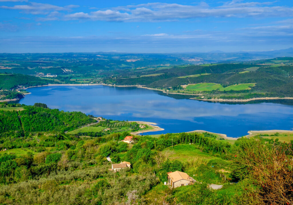 Panoramic photo from above of Lake Corbara. In the centre of the picture, there is a part of the lake located within the Tiber River Park. All around, it is surrounded by low reliefs covered in vegetation. Scattered in the lower part of the picture, the roofs of some houses can be glimpsed among the woods.