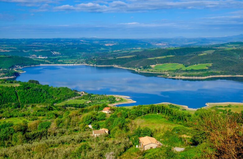 Panoramic photo from above of Lake Corbara. In the centre of the picture, there is a part of the lake located within the Tiber River Park. All around, it is surrounded by low reliefs covered in vegetation. Scattered in the lower part of the picture, the roofs of some houses can be glimpsed among the woods.