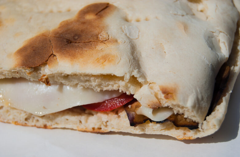 Close-up frontal shot of the hands of a lady holding the stretched out dough of torta al testo on the side edges. The dough rests on a wooden cutting board and has the typical shape of torta al testo, round and flattened.