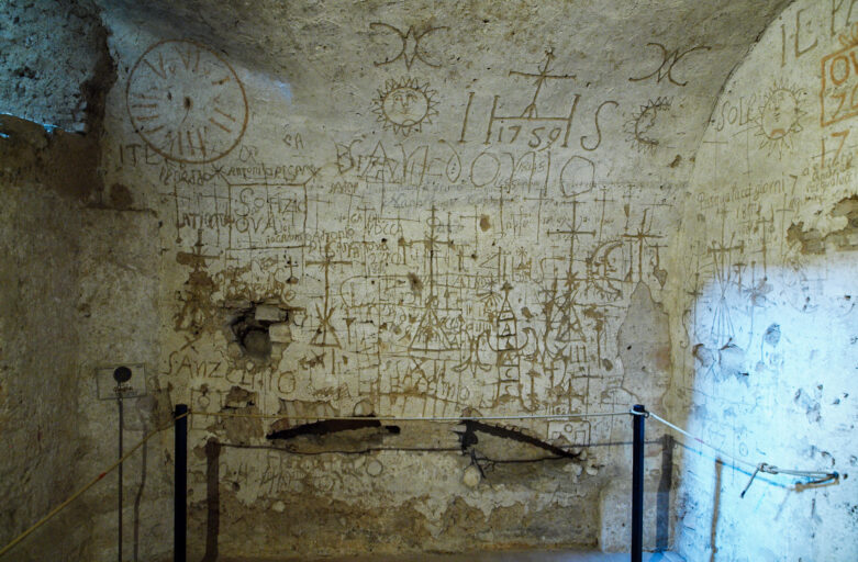 La foto raffigura le pareti interne della Sala dei Tormenti, una sala che si trova negli scavi archeologici sotterranei della città di Narni. La sala era utilizzata come stanza per gli interrogatori durante il periodo dell’Inquisizione. Le pareti sono piene di graffiti che riportano nomi, date e simboli incisi dai prigionieri che veniva interrogati al suo interno.