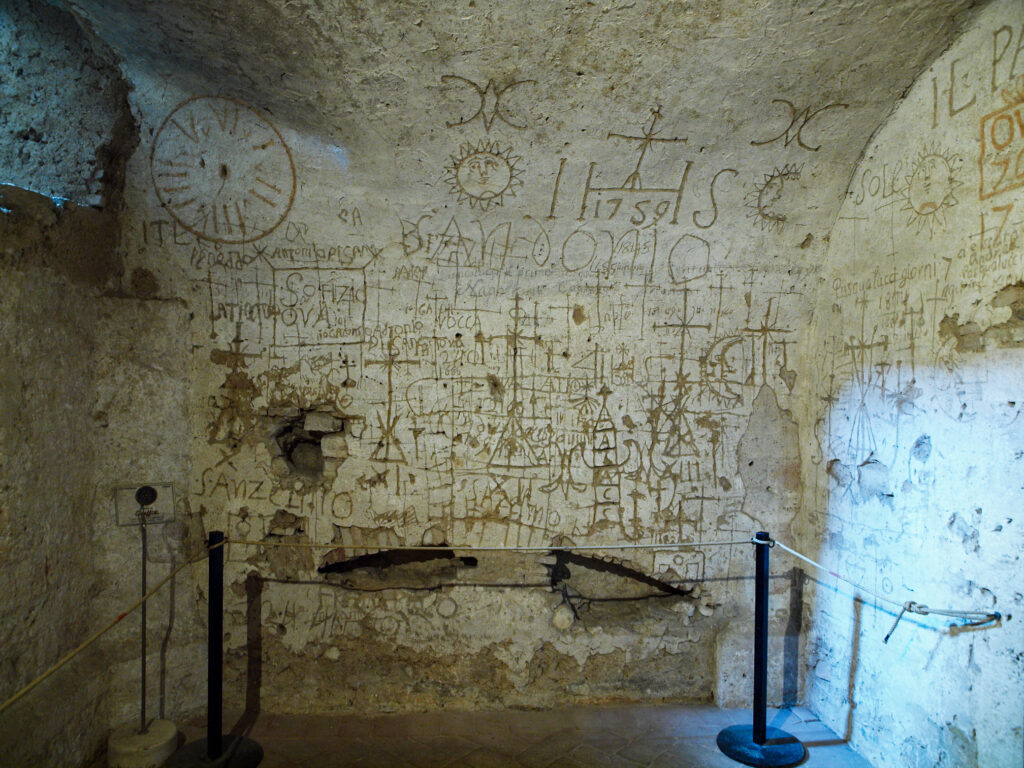 The photo depicts the internal walls of the Sala dei Tormenti, a room located in the underground archaeological excavations of the city of Narni. The room was used as an interrogation room during the Inquisition period. The walls are covered with graffiti bearing the names, dates and symbols engraved by the prisoners who were interrogated inside.