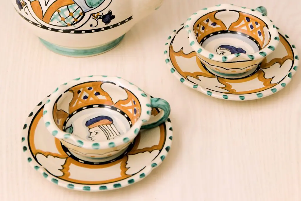 Due tazzine con piattini realizzate in ceramica di Deruta. Le tazzine e i piattini sono decorati con motivi geometrici e astratti alternati, con vari colori. Sul fondo delle tazzine sono dipinti un volto femminile e un volto maschile.