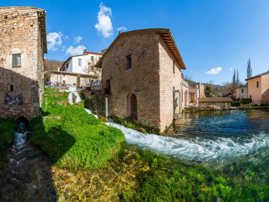 Immagine di una sezione del centro del piccolo borgo di Rasiglia. Tra i vari edifici in muratura e la vegetazione, scorre un ruscello dall’alto verso il basso e l’acqua confluisce tutta all’interno di una grande vasca collocata in basso tra gli edifici.