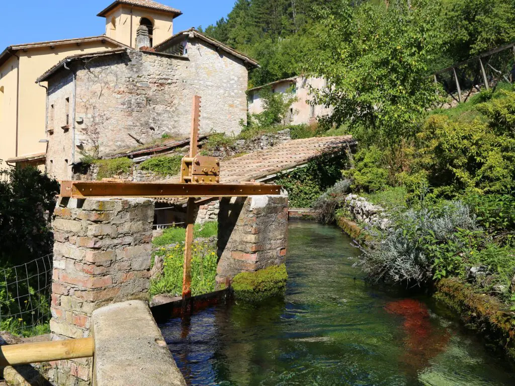 Foto di una via del centro del piccolo borgo di Rasiglia. Al centro dell’immagine, un piccolo corso d’acqua attraversa gli edifici in muratura e la vegetazione che caratterizza il paese.