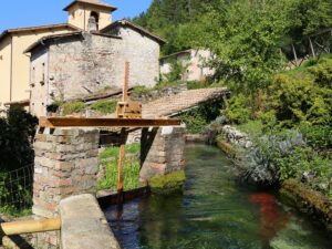 Foto di una via del centro del piccolo borgo di Rasiglia. Al centro dell’immagine, un piccolo corso d’acqua attraversa gli edifici in muratura e la vegetazione che caratterizza il paese.