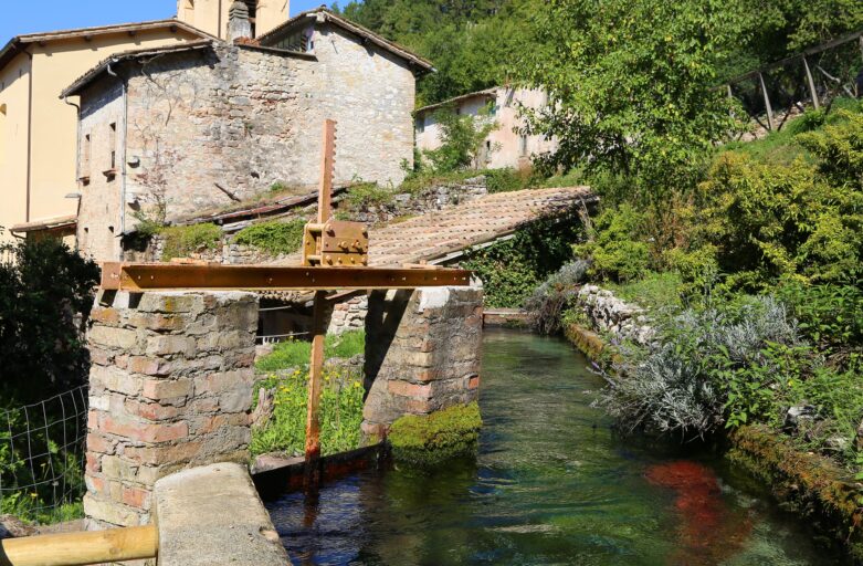 Foto di una via del centro del piccolo borgo di Rasiglia. Al centro dell’immagine, un piccolo corso d’acqua attraversa gli edifici in muratura e la vegetazione che caratterizza il paese.