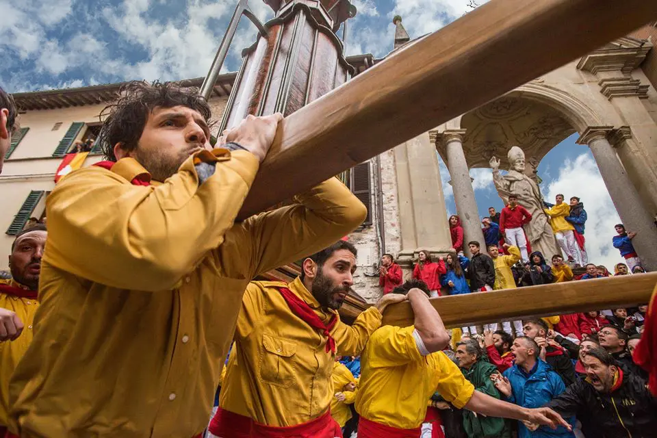 Foto che ritrae dal basso i ceraioli che stanno trasportando sulle spalle uno dei ceri di legno. Sullo sfondo, il pubblico che assiste alla corsa dei ceri in strata e dalle finestre delle case.