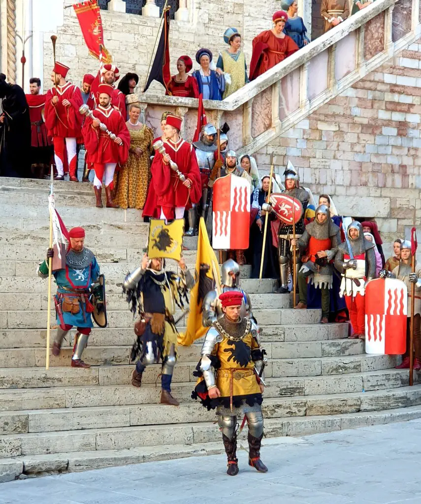 Un uomo nei panni di Braccio Fortebraccio scende dalla scalinata principale di Palazzo dei Priori durante la rievocazione storica “Perugia1416”. Ai lati della scalinata e dietro la figura di Braccio si trovano altri uomini e donne tutti con abbigliamento in stile medievale e rinascimentale, in linea con lo spirito rievocativo della manifestazione.