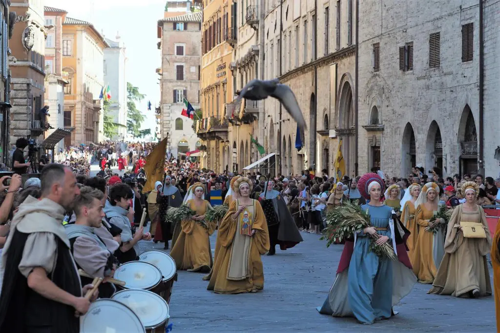 Un corteo di uomini e donne sfila per Corso Vannucci, la via principale di Perugia, in occasione della rievocazione storica “Perugia1416”. Le persone indossano abiti di epoca medievale e rinascimentale e sfilano tra la folla di cittadini e musicisti che accolgono il passaggio del corteo.