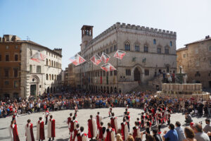 Un gruppo di sbandieratori con costumi risalenti al periodo medievale e rinascimentale tiene uno spettacolo durante la rievocazione storica “Perugia1416” al centro di Piazza IV Novembre, la piazza principale di Perugia. Gli sbandieratori sono circondati dalla folla che assiste allo spettacolo. Sulla sinistra della foto si trovano due dei principali monumeti della piazza: la Fontana Maggiore e il Palazzo dei Priori.