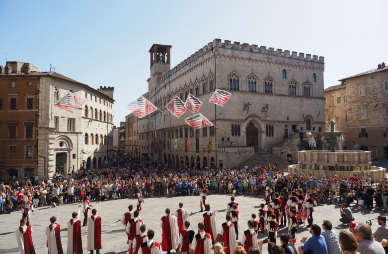 Un gruppo di sbandieratori con costumi risalenti al periodo medievale e rinascimentale tiene uno spettacolo durante la rievocazione storica “Perugia1416” al centro di Piazza IV Novembre, la piazza principale di Perugia. Gli sbandieratori sono circondati dalla folla che assiste allo spettacolo. Sulla sinistra della foto si trovano due dei principali monumeti della piazza: la Fontana Maggiore e il Palazzo dei Priori.